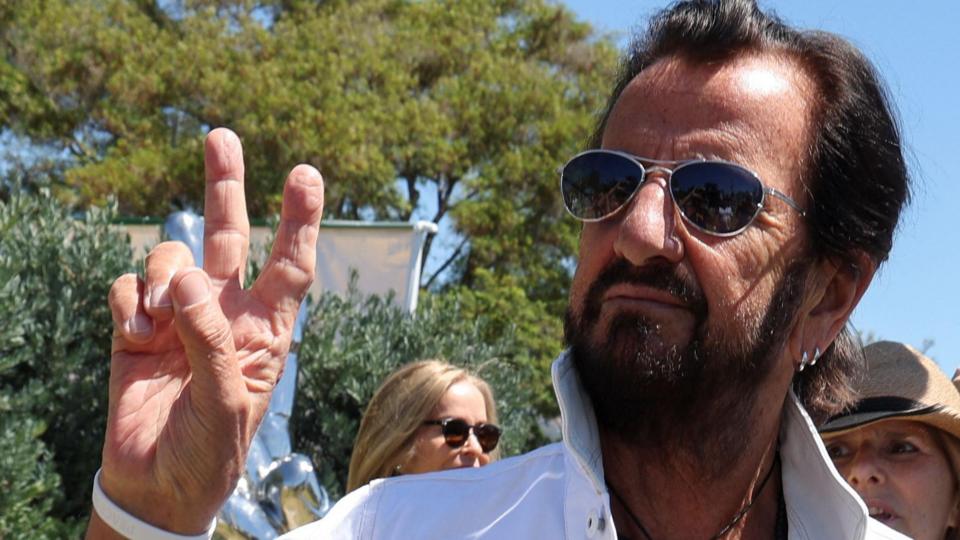 Ringo Starr, in his early 80s with black hair brushed back, a black beard and sunglasses. He is wearing a white linen shirt with the collar turned up and is holding up two fingers on his right hand in a peace symbol.