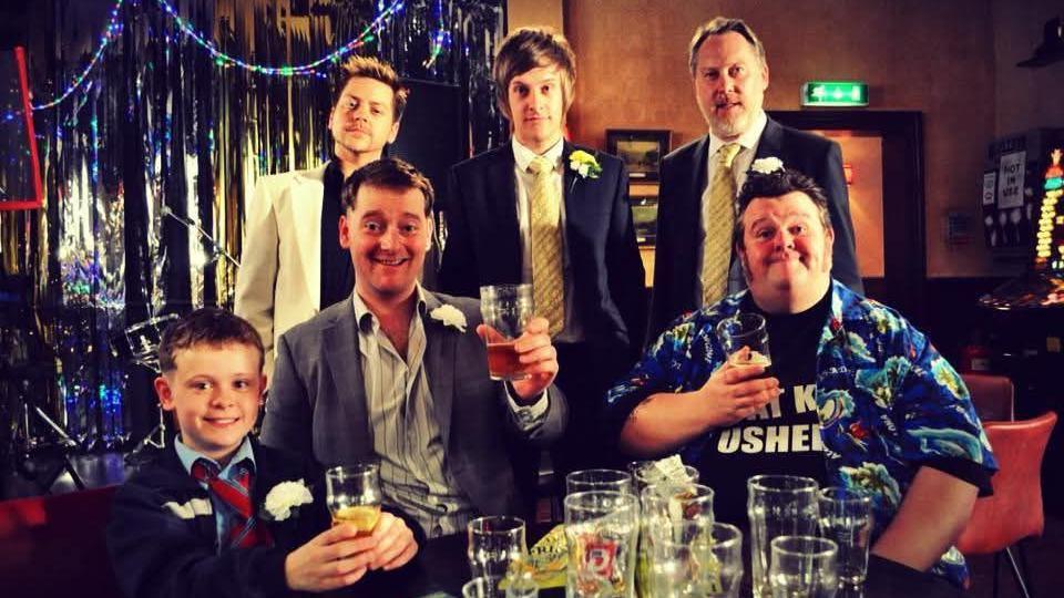 The cast of Hebburn including comedians Chris Ramsey, Vic Reeves and Steffen Peddie pose on set. Steffen is sitting at a pub table holding a pint of beer and smiling. He's wearing a brightly coloured short sleeved shirt. At the time Steffen says he weighed about 35st.