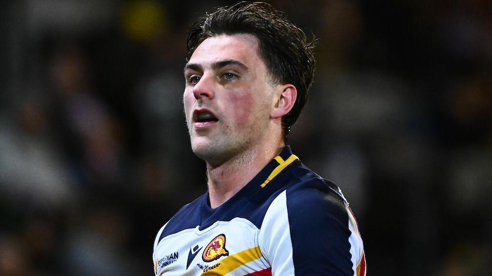 Head and shoulders of Catalans Dragons player Lewis Dodd, in white and dark blue strip with yellow and red trim.