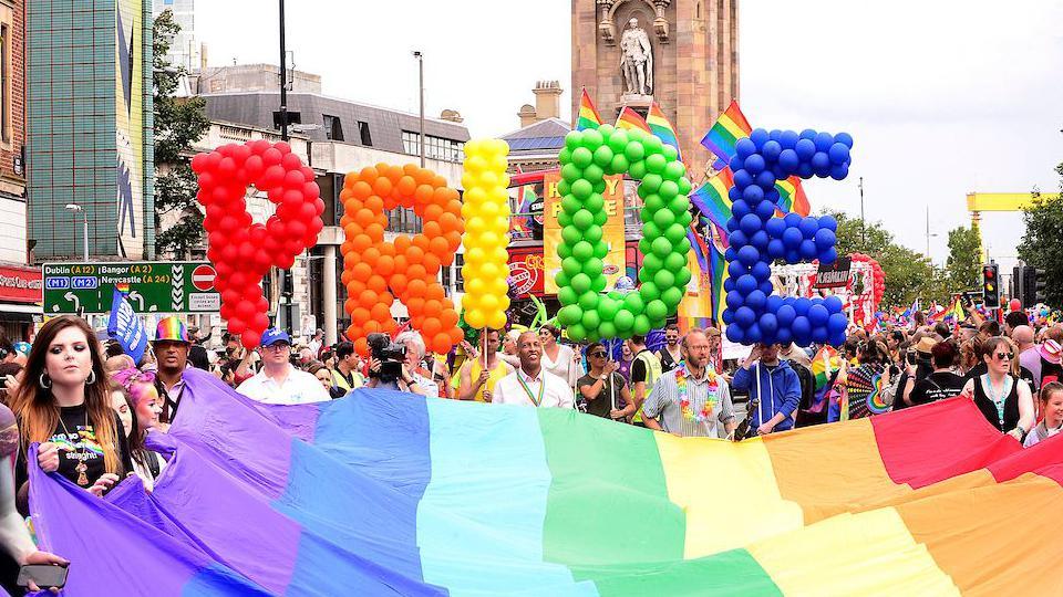 Large balloon letters that say Pride in rainbow colours held above a large rainbow flag.