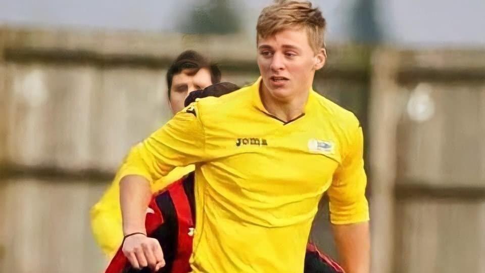 A teenage boy with short blonde hair and wearing a yellow football shirt, looks to the right as he runs on a football pitch. Two other players are slightly visible behind him.