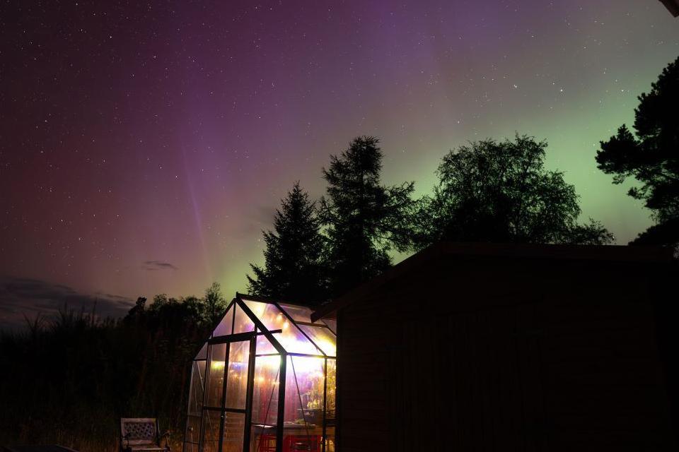 A greenhouse lit up by lights and aurora borealis in the night sky above