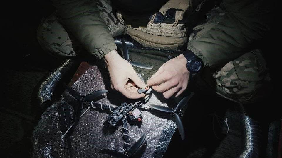 A soldier fixing or setting up a black drone in the dark. Only his hands and knees are visible, lit up by what seems to be a head torch shining down from above.