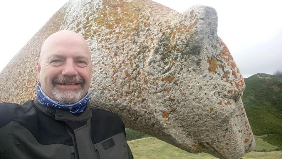 A selfie of John Wood who is wearing a black motorcycle jacket. He is smiling at the camera and posing in front of a statue. 