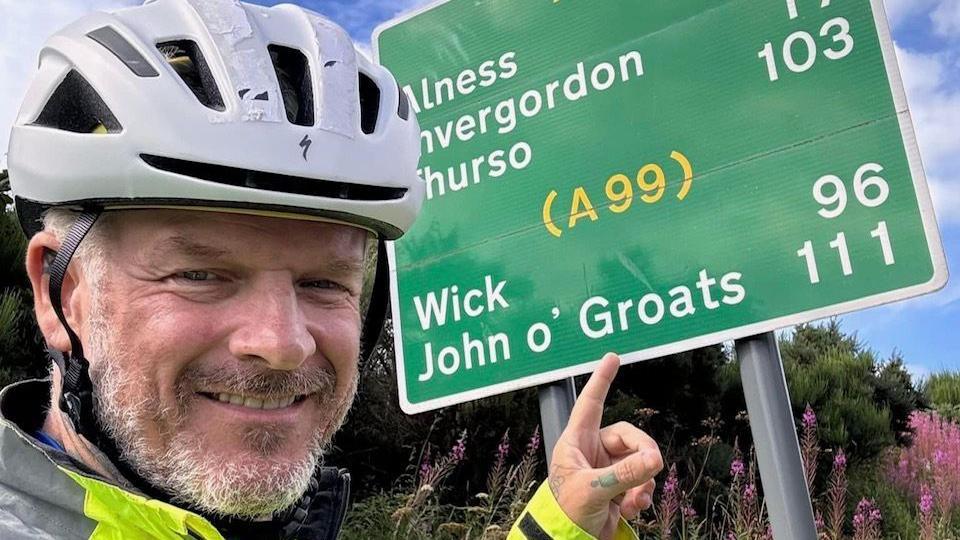 Mathew Pritchard pointing to a sign that says John O' Groats 111. He is wearing a cycling helmet and reflective yellow coat.