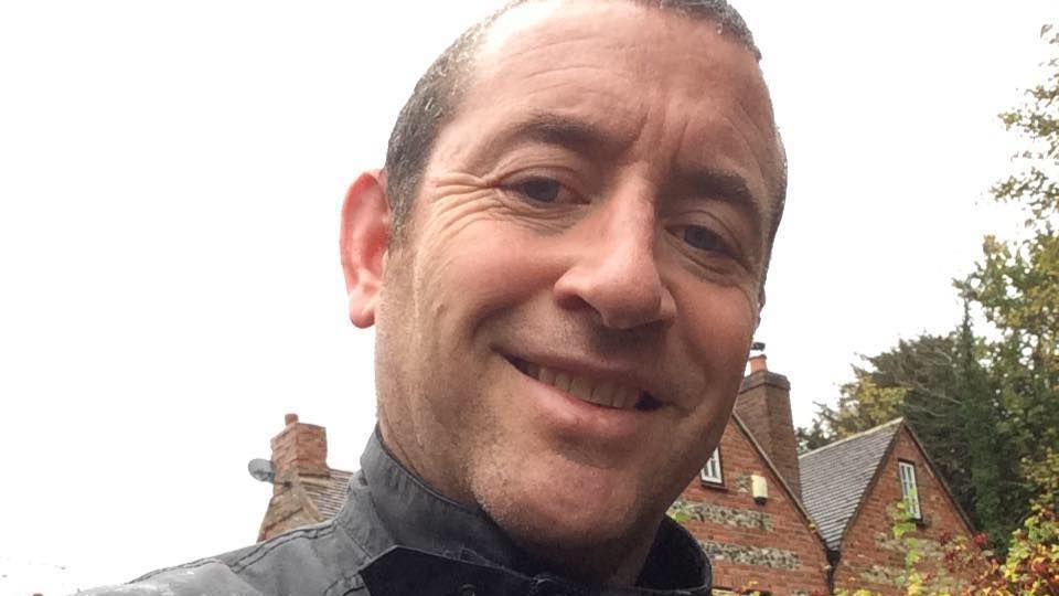 A selfie taken by David Carrick, who has short dark hair. The black collars of his jacket are pictured and a house is behind him. He is smiling.