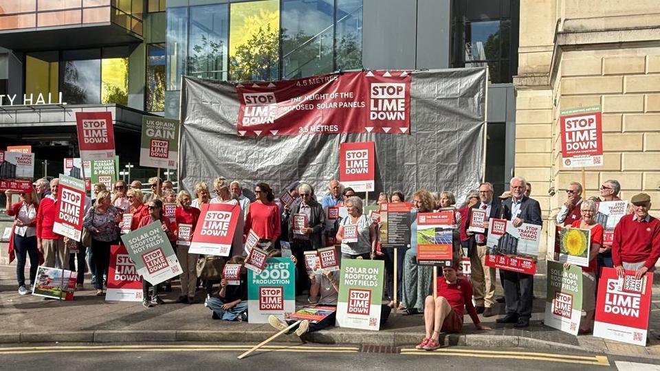 A large group of protesters outside the Wiltshire Council building in Trowbridge. There is a large banner behind them and many of the protesters have their own smaller banners and placards that read: Stop Lime Down.
