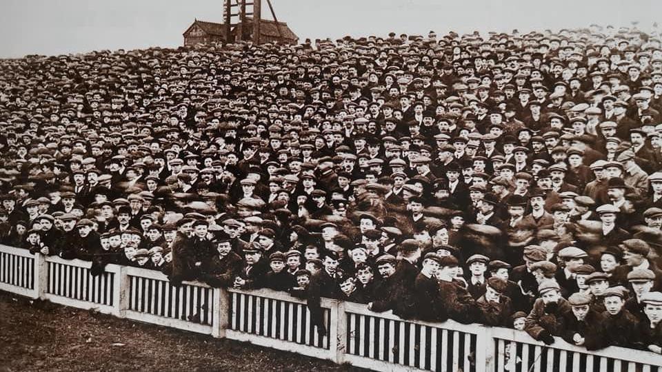 A black and white photo of the Spion Kop. Hundreds of football fans, many wearing flat caps, stand behind a wooden fence