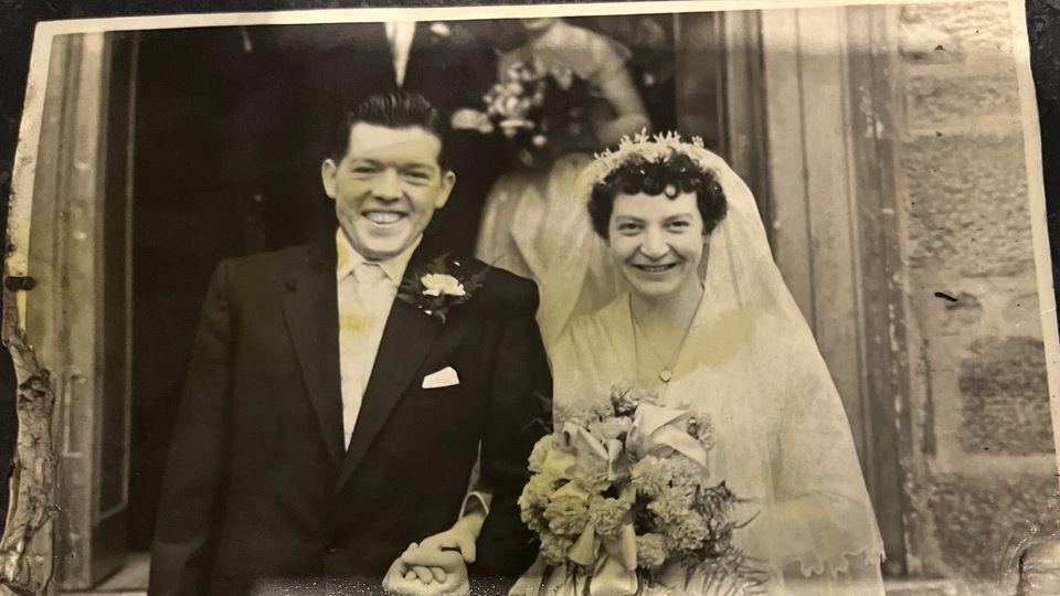 A black and white picture of a couple on their wedding day outside a church. The man wears a black suit and has dark hair and holds the woman's hand. The woman wears a white dress and a veil and holds a bouquet of flowers. 