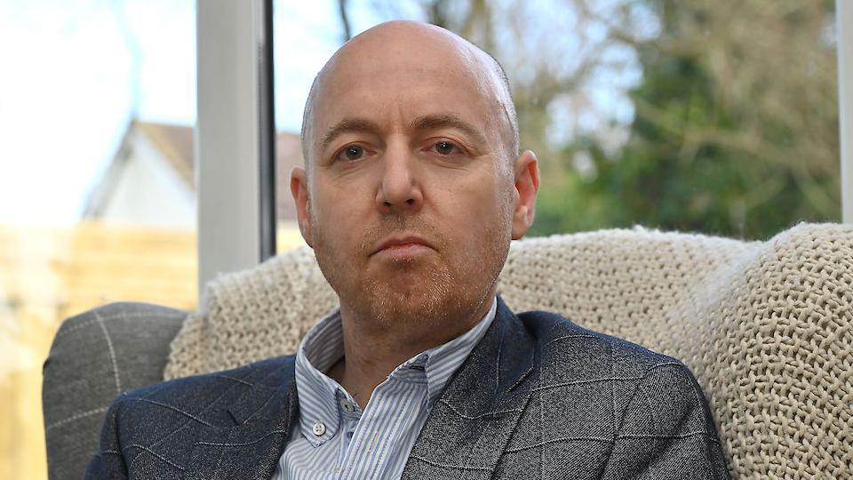Jon Burrows is in a grey chequered suit and blue and white striped shirt.
He is sitting on a grey arm chair with white blanket draped over it.