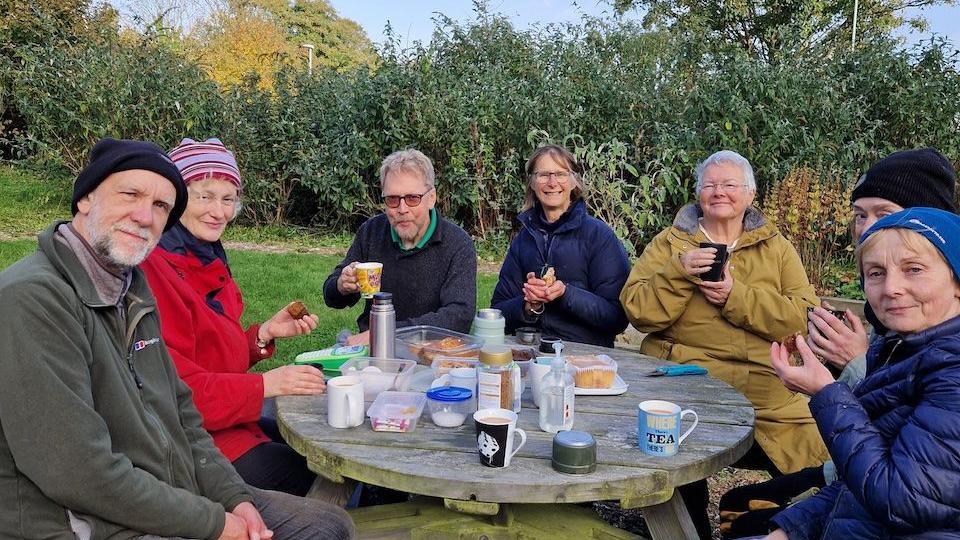 Seven men and women sit around a round picnic table in outdoor coats as they drink tea, and eat snacks with the bushes from the garden in the background.