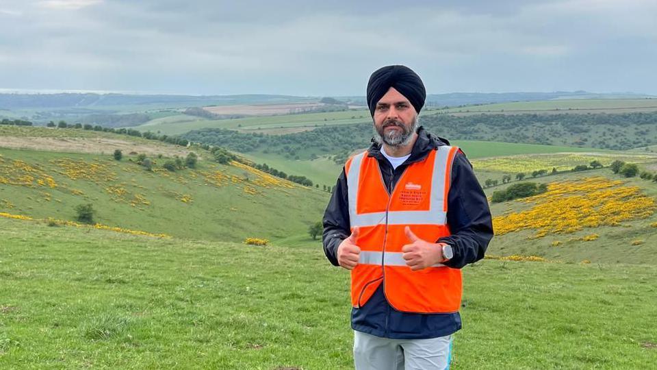 A man wearing an orange high-vis vest with his thumbs up. He is stood at the top of a series of grassy hills