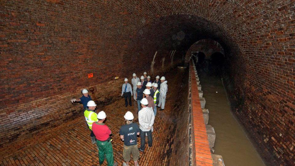 A group of people in white hard hats inside a brown brick sewer chamber