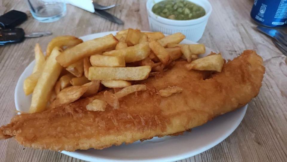 A plate of fish and chips with a pot of mushy peas in a pot beside it, with cutlery on the table.