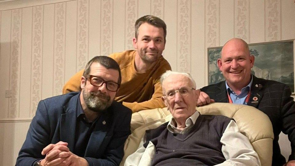 An elderly gentleman, Jack Mortimer, is sitting in a chair and is surrounded by some of the JLR marathon runners on his birthday. He is wearing a cream shirt and a brown pullover.