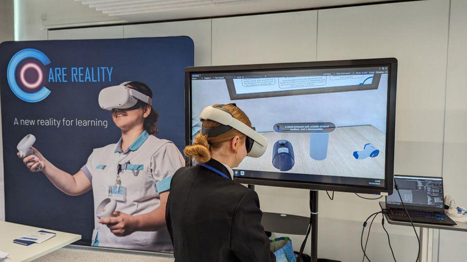 A woman, with her hair tied up, stood with a white VR set on in front of a screen. There is a blue sign on the left with an image of a woman with a VR headset on.