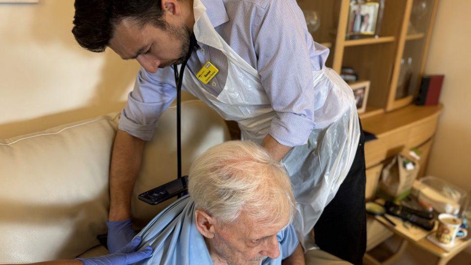 A doctor leans over a patient who is on a sofa. A cupboard is on the right of the photo.