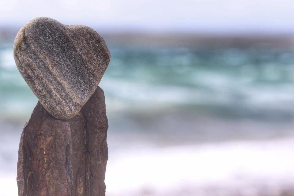 A small "heart shaped" stone perched on top of another stone with rough waters out of focus in the background