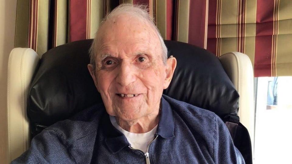 An elderly man smiling while sitting at a table, with striped curtains in the background.