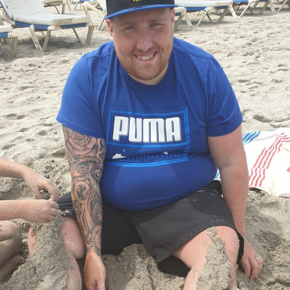 A man in a hat sits on the beach while sand is piled on his legs