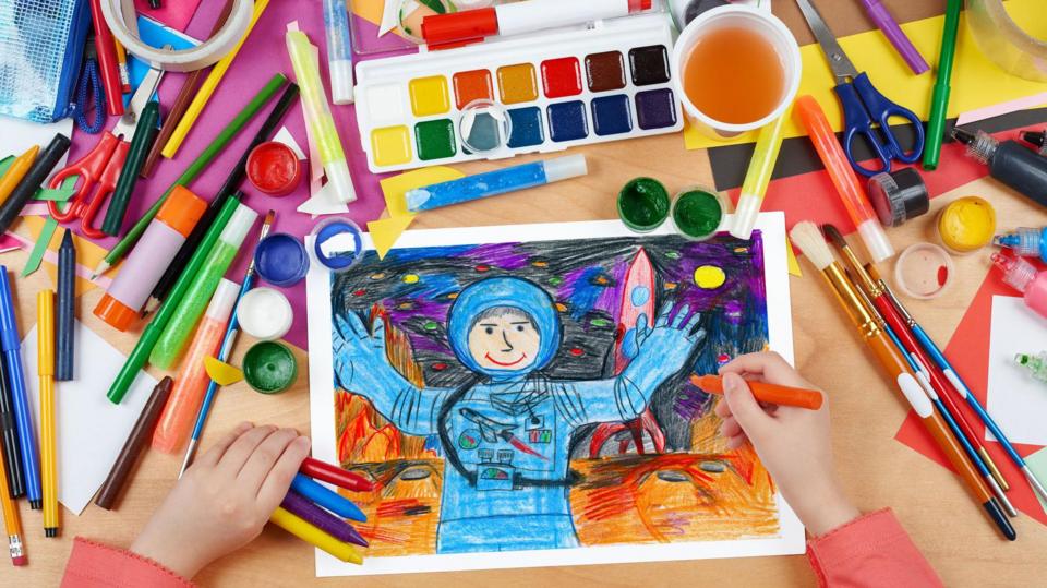 seen from above - child's hands drawing an astronaut in space in bright coloured pencils, surrounded by stationery