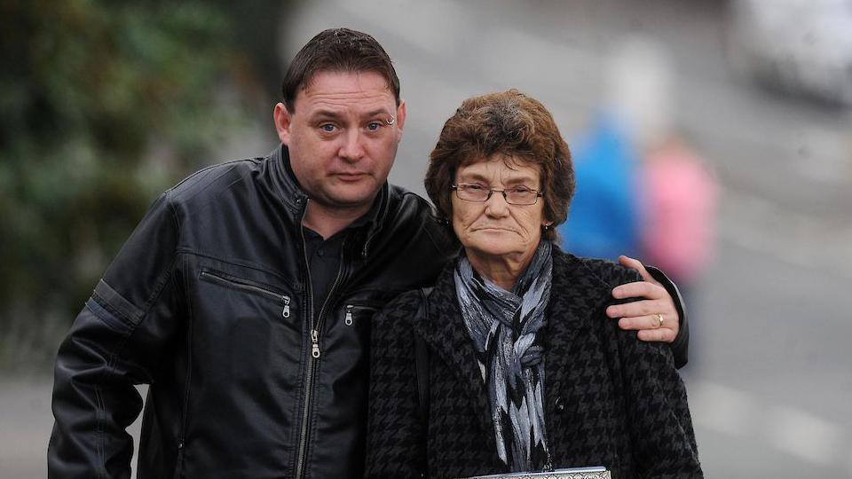 A man in a black leather jacket brown hair with his arm around a woman with a black jacket on and grey scarf. She has brown short hair and glasses. The background is blurred. They are looking at the camera.