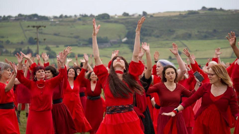 A group of women dressed in red dressed dance on the moors, many with their hands in the air, as part of an event to celebrate Kate Bush