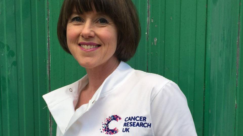 Caroline Jones on 31 December 2015. She has brown hair which falls just below her ears and is smiling at the camera. She is wearing a white Cancer Research UK lab coat with the charity's logo on it.