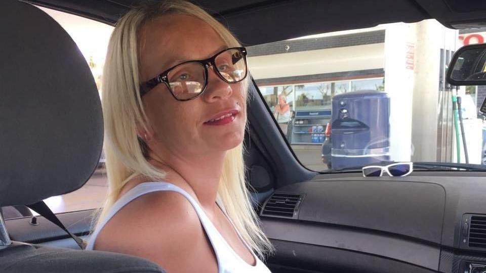 A lady sat in the front passenger seat of a car. She has blonde hair and is wearing a white vest top. She has black glasses. The car is parked at a petrol station. There are some white sun glasses on the dashboard. 