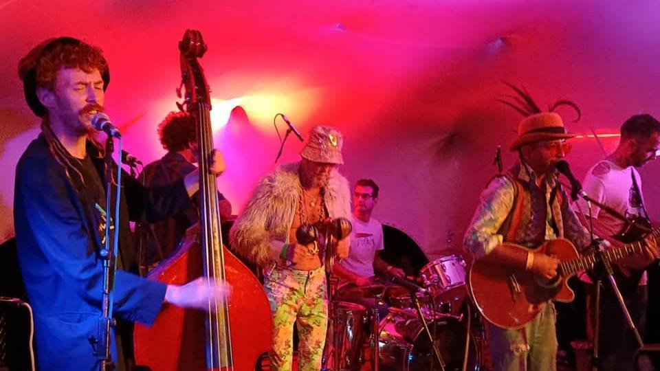 Opaque playing a gig with instruments including a cello, maracas, drums and guitar. There is a magenta light being cast on them making the scene look warm. Some of band are also wearing unusual outfits with one member having a glittery bucket hat, a fluffy jacket and flowery trousers. Another wears a hat with feathers on.