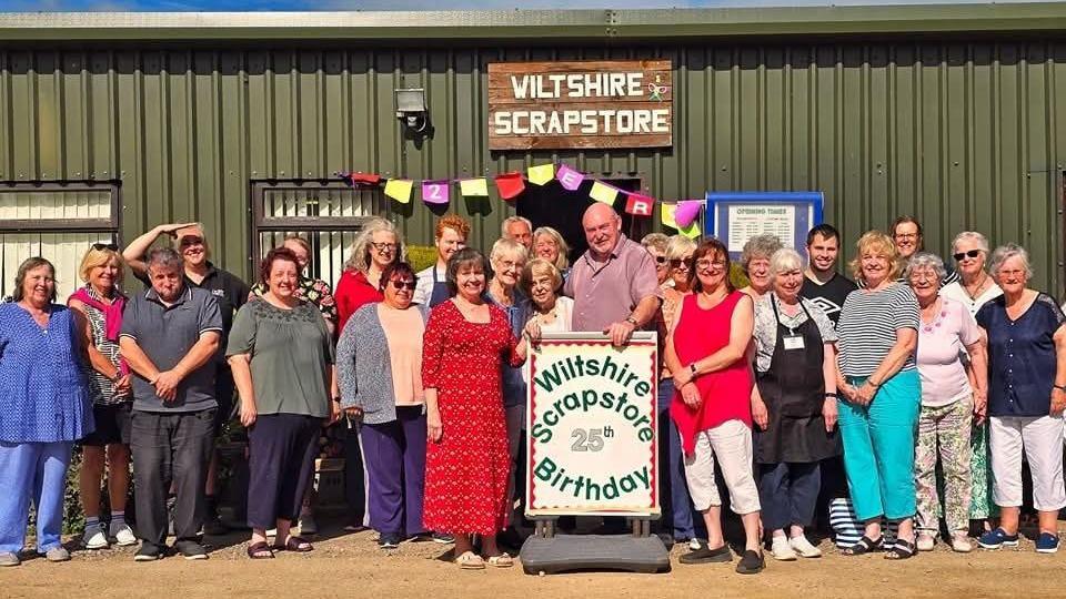 A group photograph outside the Wiltshire Scrapstore warehouse. More than 20 adults stand together in the sunshine with an information board in front reading "Wiltshire Scrapstore 25th birthday".