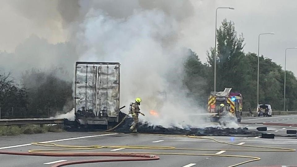 A lorry which has been on fire. There are still some flames coming from the lorry and smoke is billowing into the sky. Firefighters are tackling the blaze with hoses and a fire engine is parked near the vehicle. Debris of tyres is spread across the road. 