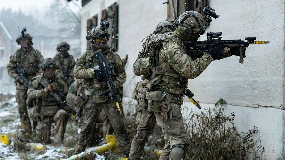 British Army officer cadets from the Royal Military Academy prepare to assault a building during an exercise in Germany. They wear combat uniforms and wield guns as snow falls around them.