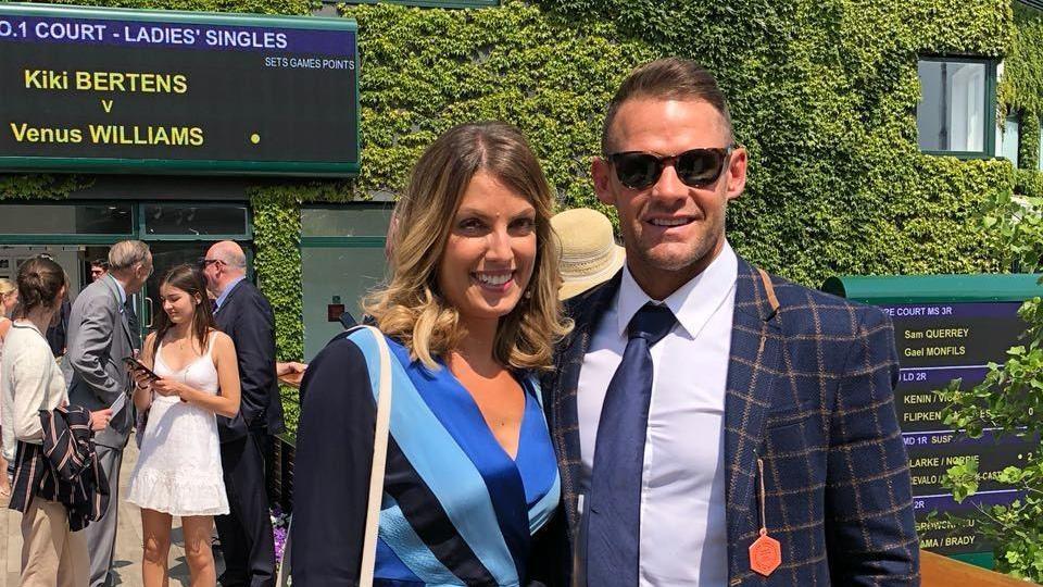 Andrea, dressed in a blue dress, stands next to Lee, wearing a blue suit, smiling at Wimbledon 