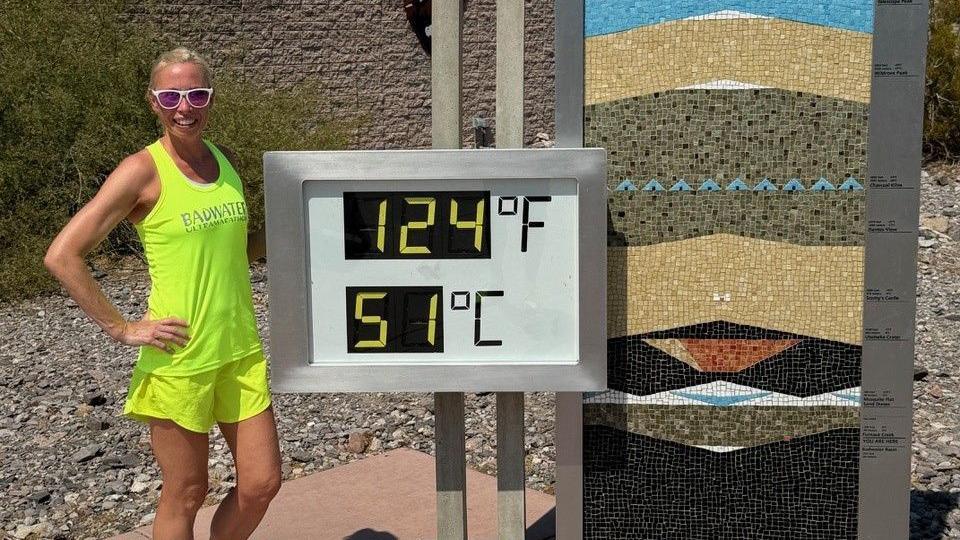 A woman, Laura Watts, is wearing a yellow vest and shorts and sunglasses. She has tied back blonde hair and is smiling at the camera. She is standing to the left of a white sign with grey frame with the digits 124F written above 51C