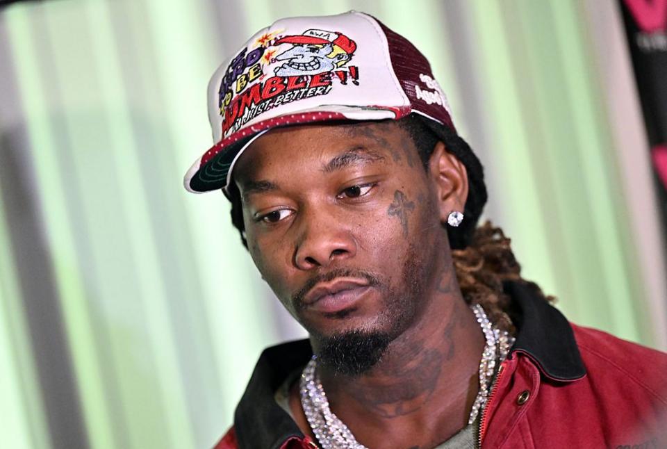 Offset is a man, he has brown eyes and is wearing a white and red cap.
He has a diamond stud earring and is wearing a large silver chain. He is wearing a red jacket with a black collar. He has a tattoo on his cheek below his eye of a cross and tattoos on his neck.