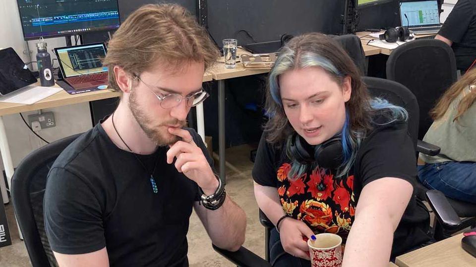 Image shows 23 year old Iz Head wearing a printed black and red tee shirt in conversation with a male colleague, wearing a plain black tee shirt