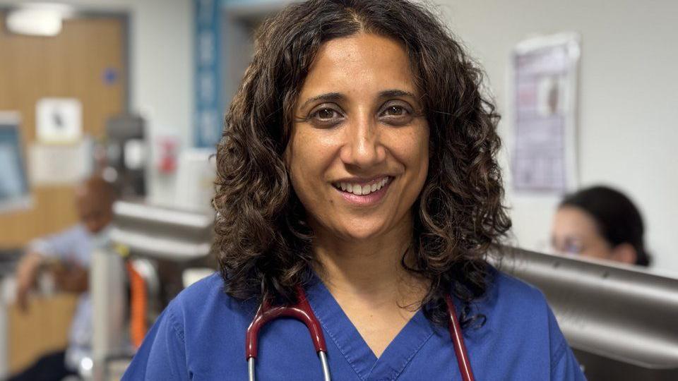 A doctor with dark hair and in blue uniform is smiling. Two people in the background are blurred.