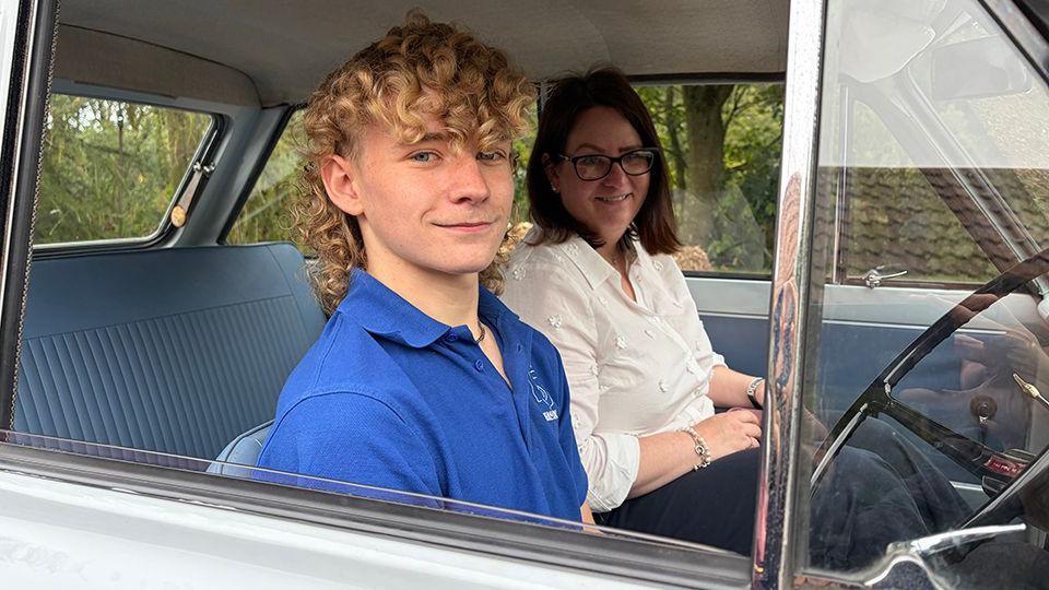 Lathan is sat in the driver's seat of the car and is smiling at the cancer. Anona is sat next to him.