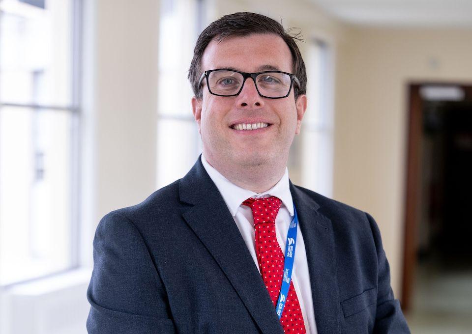 Chief operating officer, Simon Illingworth looking at the camera. The picture shows his head and shoulders and he is wearing black glasses. He has a red tie and a black hacket on. He is smiling and standing in a hospital corridor