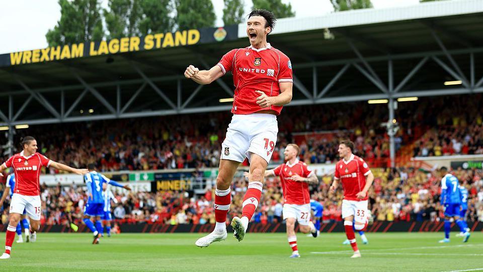 Wearing a red home Wrexham kit, Kieffer Moore jumps in the air to celebrate scoring for Wrexham against Sheffield Wednesday at the Cae Ras. The Wrexham Lager Stand is in the background. In the background, Lewis O'Brien, Nathan Broadhead and George Dobson run towards Moore. 