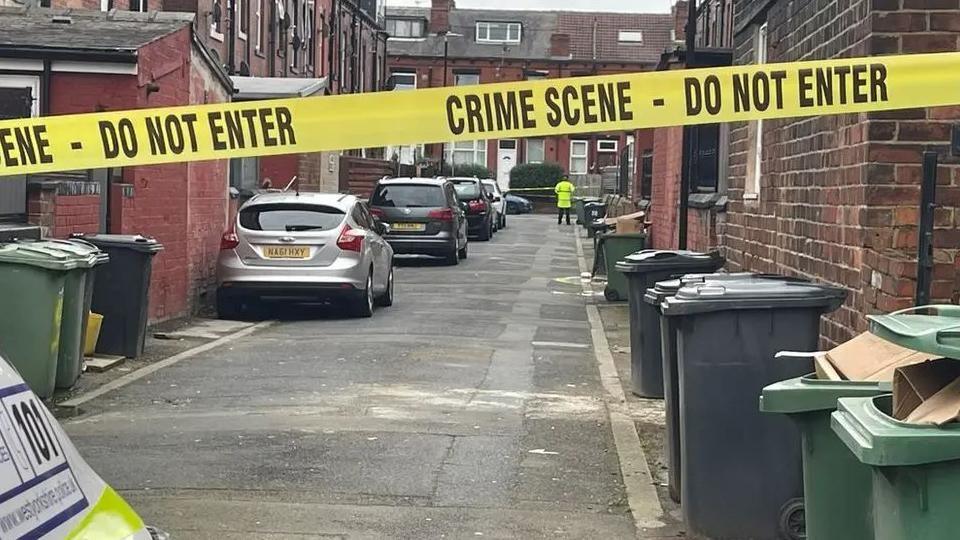 An alleyway with cars and wheelie bins in, and police tape across it