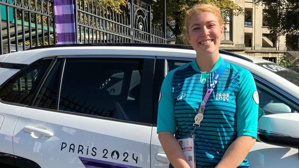 A woman stands in front of a white car with "Paris 2024" branding on its side. She's smiling and wearing an official Paris 2024 top - it's bright blue with black horizontal stripes across the torso. She's also wearing a lanyard with an official ID badge hanging off it.