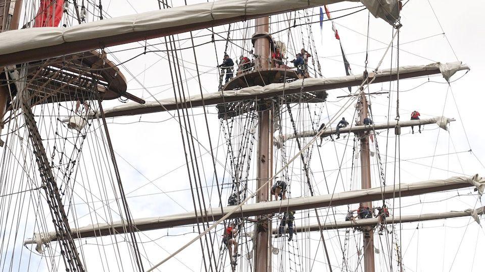 Masts and riggin of a tallship with people wearing harnesses high up in the air, working on the sails