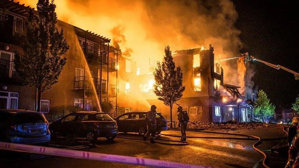 Two firefighters can bee seen silhouetted in a car park while in the background a building is well alight and water jets can be seen targeting the blaze