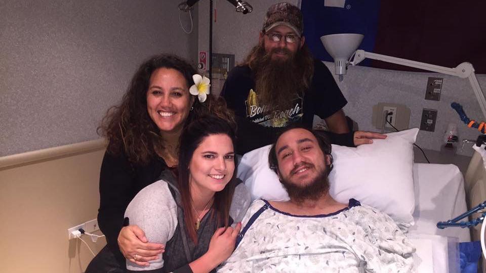 Noland lying in a hospital bed. He and his family members are all smiling broadly. They all look very happy.