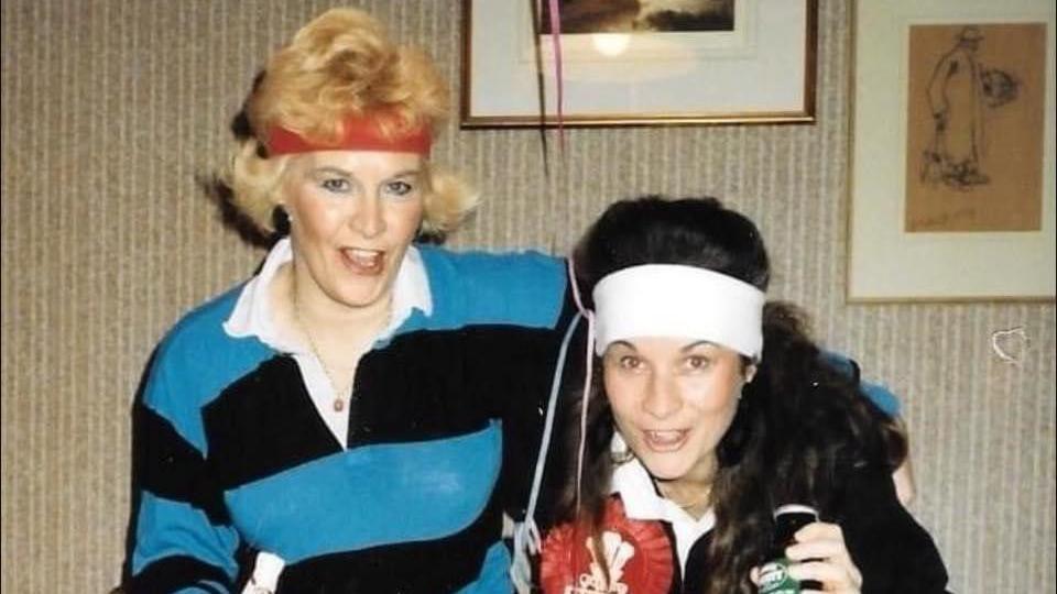 An older woman wears a red sweatband with a black and blue dress holding her younger daughter who wears a white sweatband, both are animated and a laughing towards the camera