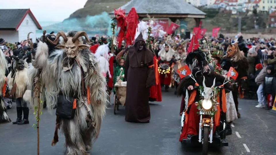 People dressed up as a mythical horned beast called Krampus walk in a parade through Whitby, holding red and white flags. One person is riding a motorbike. 