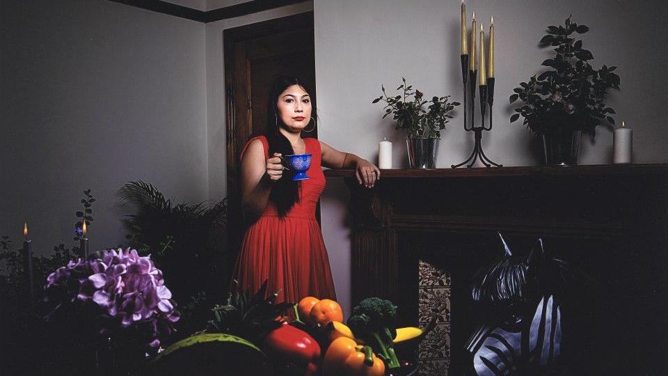 Ffoto o fenyw mewn ffrog goch yn sefyllfa ar bwys lle tan. Mae lliwiau cryf yn y llun - coch y ffrog, coch a melyn ac oren a gwyrdd mewn basged o ffrwythau a llysiau ym mlaen y llun, a blodau piws ar yr ochr chwith. 