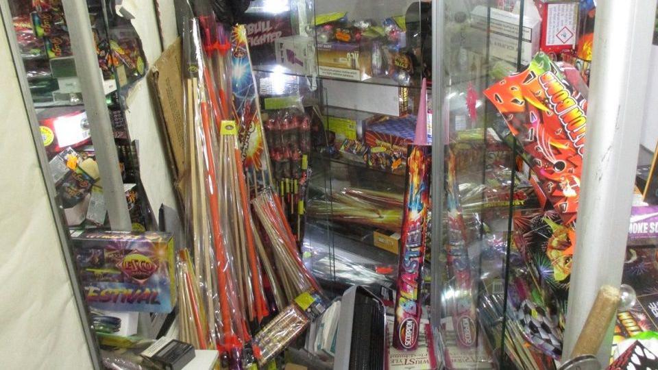 A collection of fireworks lumped together in what looks like a storage room. They are an array of different colours and are mostly a long wooden shape.